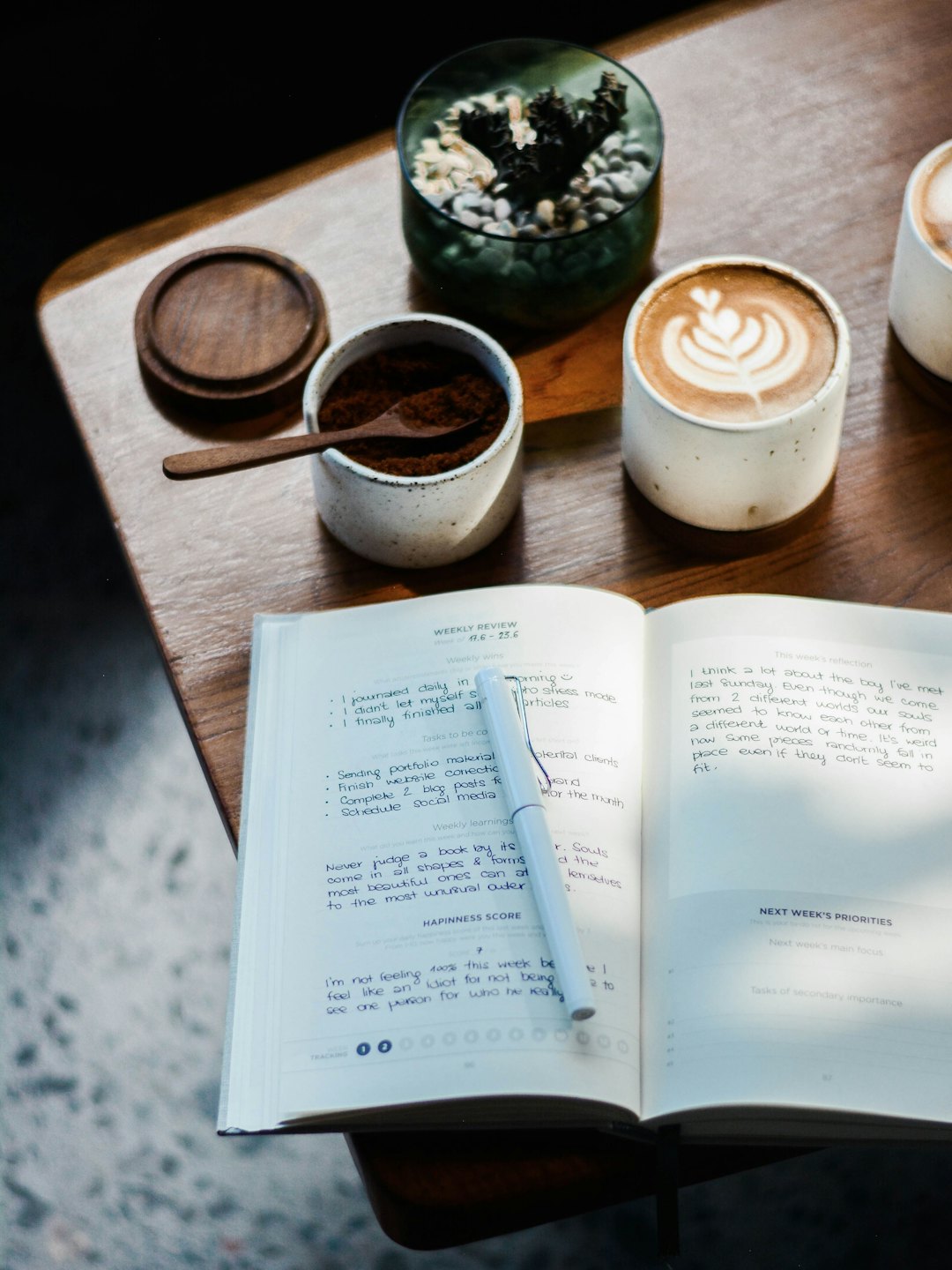 Journal with pen, coffee, and plant on wooden table.