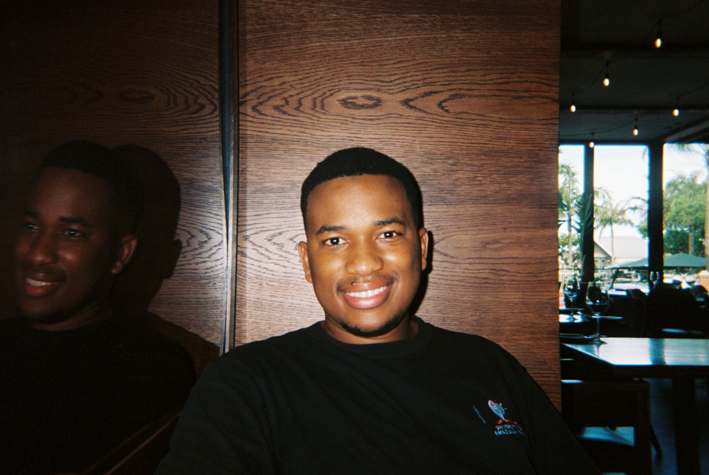 A man in a black t-shirt smiles indoors near wooden paneling.